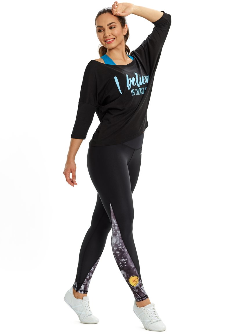Fekete oversized ing kék felirattal és háromnegyedes ujjakkal, fekete leggings-szel párosítva, amelyek színes virágmintásak az aljukon. Fehér sportcipő.