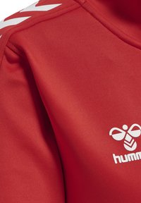 Rødt stof med hvid Hummel-bi-logo og chevron-design på skulderen, sandsynligvis en del af et sportstøj.