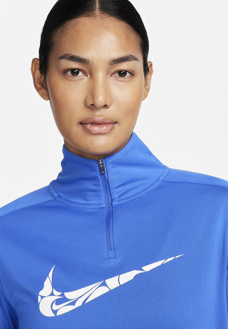 Mujer con el cabello oscuro recogido que lleva una sudadera azul con cremallera que presenta un logo de Nike en forma de swoosh blanco en el pecho.