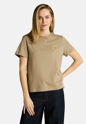 Blondynka w beżowej koszulce z małą grafiką cytryny i napisem „Limon”, w połączeniu z ciemnoniebieskimi jeansami, stojąca na białym tle.