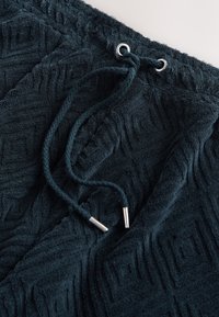 Marinblå frottétyg med ett strukturerat geometriskt mönster. Har dragsko med silverfärgade metalländar och dekorativa sydda detaljer.