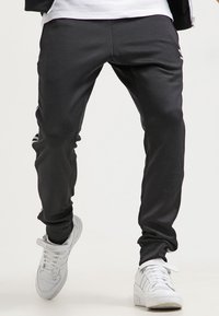 Svarta tajta sweatpants med en slät textur, med tre vita ränder längs sidorna och en logotyp framtill. Bärs med vita sneakers.