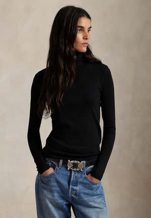Kvinne med langt mørkt hår iført en svart turtleneck og blå jeans, hender i lommer, ser mot høyre mot en ensfarget bakgrunn.