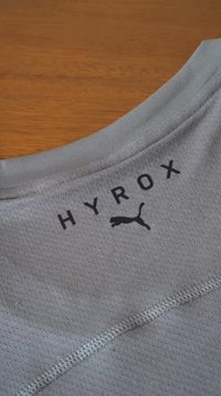 Γκρι ύφασμα πλέγματος με τη λέξη "HYROX" και μαύρο λογότυπο Puma τυπωμένο κοντά σε μια ραφή σε ξύλινη επιφάνεια.