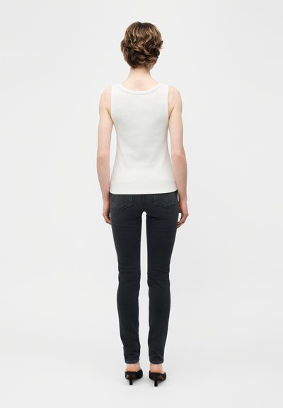 Personne aux cheveux courts et bouclés, vue de dos, portant un haut blanc sans manches, un jean skinny noir et des talons noirs sur un fond blanc.