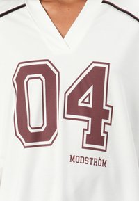 Weißes Trikot mit V-Ausschnitt, das große bordeauxrote Zahlen "04" in weißem Rand zeigt, und "MODSTRÖM" darunter in passender bordeauxroter Schrift aufgedruckt.