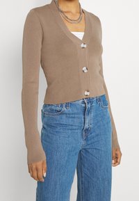 Kvinna som bär en figursydd ljusbrun V-ringad kofta med tre knappar över en vit topp och högmidjade blå jeans.