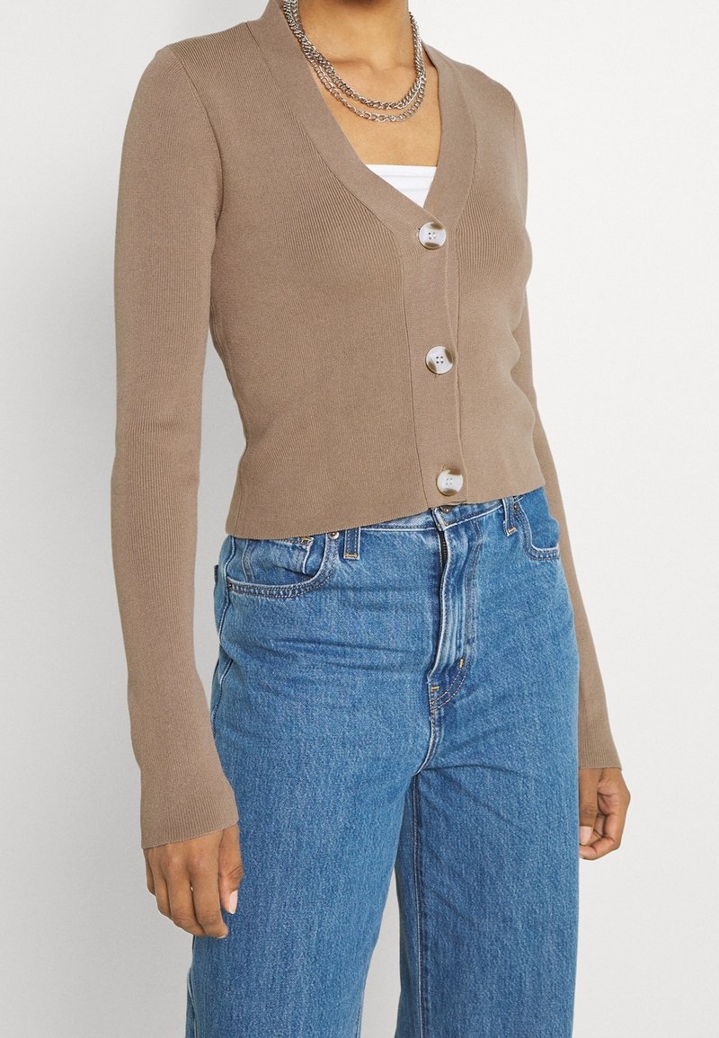 Kvinna som bär en figursydd ljusbrun V-ringad kofta med tre knappar över en vit topp och högmidjade blå jeans.