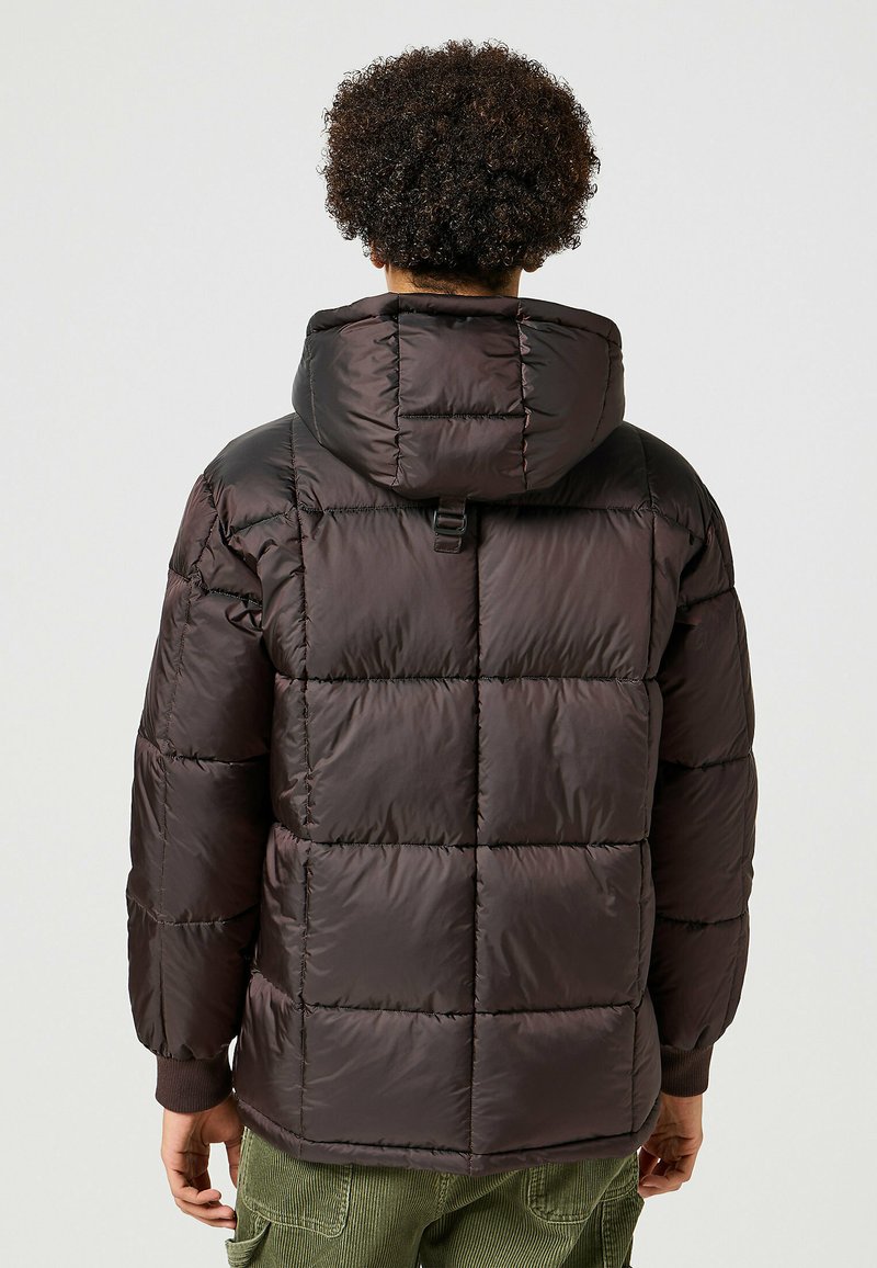 Giacca puffer di colore marrone scuro, con design trapuntato, cappuccio e polsini a costine. Il materiale appare lucido e ha una consistenza morbida.