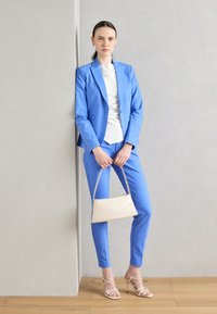 Světle modrý oblek na míru, zahrnující sako a úzké kalhoty, doplněný bílým topem a krémovou kabelkou. Look doplňují béžové sandály na vysokém podpatku.
