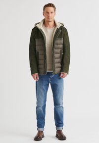 Grüne Pufferjacke mit einem mit Schaffell gefütterten Kapuze, mit gesteppten Abschnitten und Seitentaschen, kombiniert mit hellblauen Jeans und dunklen Stiefeln.