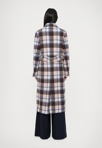 Manteau à carreaux en beige, marron et bleu. Fait de laine, il dispose d'un col, d'un détail de ceinture au dos et de poignets à boutons. Porté par-dessus un pantalon sombre.