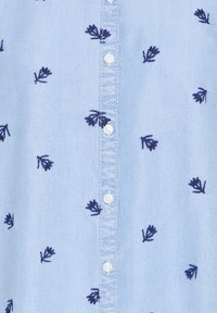 Lyseblå skjorte med hvite knapper og små mørkeblå broderte bladmønstre spredt over stoffet.