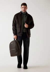 Veste marron zippée avec poche poitrine, col roulé gris, jean foncé et un sac à dos marron texturé avec une fermeture éclair à l'avant. Chaussures noires.