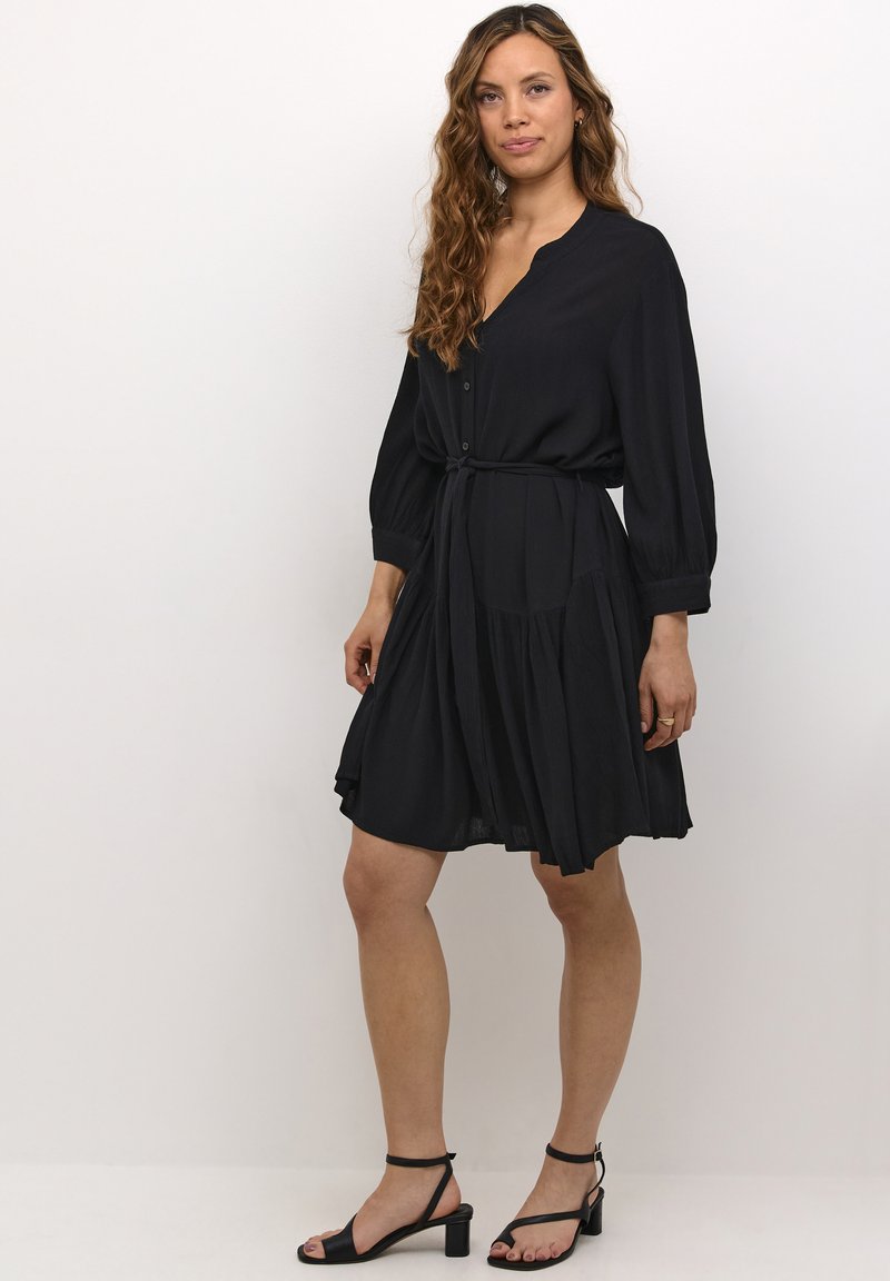 Schwarzes Hemdkleid mit langen Ärmeln, geraffter Taille und knie-langer Saum. Hergestellt aus weichem Stoff mit einer leichten, fließenden Textur.