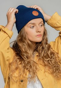 Niebieska dziana czapka z okrągłą brązową skórzaną łatą. Model ma na sobie żółtą kurtkę. Włosy są długie i faliste. Tło jest neutralne.