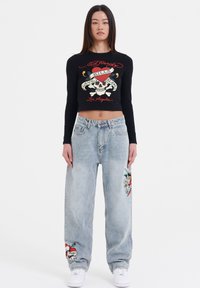 Black long-sleeve cropped top with graphic skull design, paired with light blue, loose-fit jeans featuring colorful floral embroidery and white sneakers.