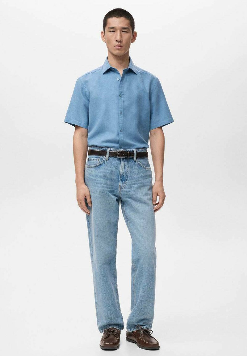 Hellblaues Kurzarmhemd; vorne mit Knöpfen. Hellblaue Jeans mit geradem Bein. Braune Schuhe mit abgerundetem Zehenbereich. Dunkler Gürtel. Schlichter Hintergrund.