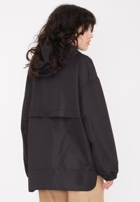Veste noire surdimensionnée avec capuche, texture lisse et poignets élastiques. Présente un design de dos superposé et une coupe ample.