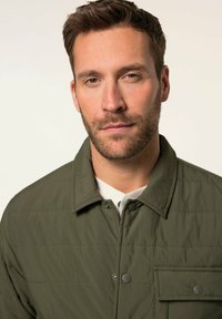 Grüne gesteppte Jacke mit Kragen, vorderen Druckknöpfen und einer Brusttasche. Der Stoff wirkt weich mit einer glatten Textur.