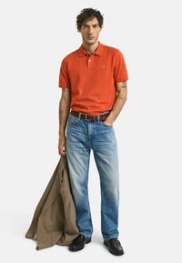 Rød polo-shirt med korte ærmer, blå jeans og sorte sko. Holder en brun mønstrede blazer. Casual pasform og afslappet design.