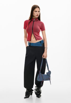 Vrouw draagt een rood, korsetachtig crop top met korte mouwen en ritssluiting, zwarte wijde broek met een opgevouwen denimriem, zwarte laarzen en houdt een blauw denim handtasje vast.