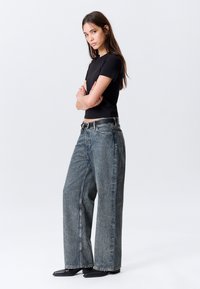 Μαύρο εφαρμοστό μπλουζάκι συνδυασμένο με φαρδιά, ξεθωριασμένα μπλε τζιν με λεπτή υφή και θηλιές για ζώνη. Το μοντέλο φορεί μαύρα υποδήματα.