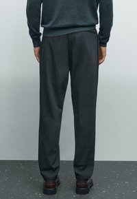 Man wearing black trousers, dark sweater, and brown shoes standing on dark speckled floor against gray wall, viewed from behind.