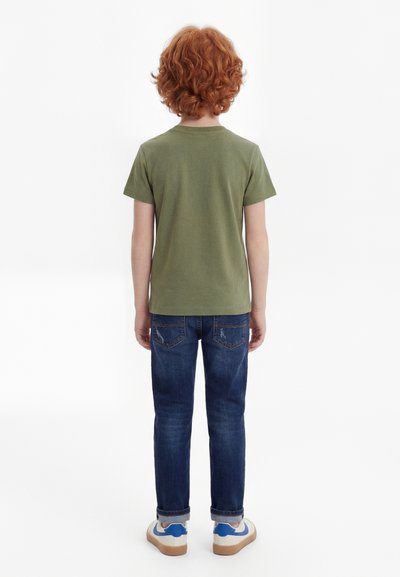 Göndör vörös hajú gyermek olívazöld pólóban, sötétkék farmerben és fehér tornacipőben, háttal állva fehér háttér előtt.