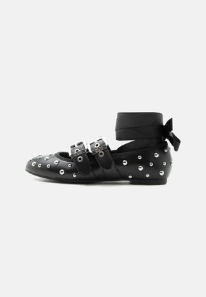 Black flat shoe with silver studs, double buckle straps, and a wrap-around ankle ribbon tied in a bow.