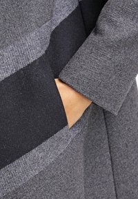 Abrigo de mezcla de lana gris y negro con un patrón a rayas, que presenta un diseño moderno y un gran bolsillo externo en el lateral.