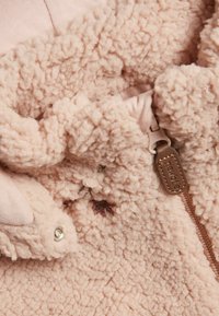 Mjuk beige fleece-tyg med en dragkedjesnodd märkt "newbie" och små tryckknappar på ett texturerat material.