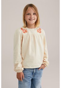 Blusa crema con tessuto testurizzato, ricamo floreale arancione sul collo, maniche arricciate e scollatura rotonda. Abbinata a jeans in denim blu.