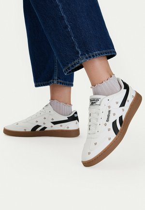 Bílé tenisky Reebok s černými pruhy a malými vzory včel, nošené s světlými ponožkami a tmavě modrými zkrácenými džínami.