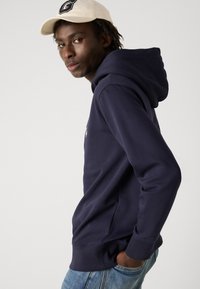 Felpa blu navy con tasca a marsupio e polsini a costine, indossata con jeans azzurri chiari. Un cappellino da baseball crème aggiunge contrasto.