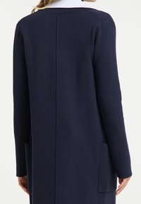 Langer navyblauer Cardigan aus weichem Strickstoff. Er verfügt über einen geraden Schnitt, zwei Fronttaschen und einen Kragen mit einer kontrastierenden Farbe am Hals.