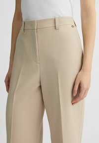 Pantaloni beige su misura realizzati in un tessuto liscio, con vita dotata di passanti per cintura e un piccolo logo in metallo.