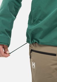 Veste imperméable verte avec cordon de serrage ajustable à l'ourlet ; pantalon beige avec une poche zippée et un détail de logo. Accent sur la fonctionnalité et les matériaux.