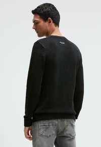 Mann trägt schwarzen Rundhalsausschnitt-Pullover und ausgewaschene graue Jeans, steht leicht seitlich gedreht und zeigt das hintere Etikett am oberen Rücken des Pullovers.