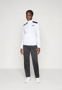 Biała kurtka na zamek z czarnymi akcentami na ramionach i logo, połączona z ciemnoszarymi spodniami i białymi sneakersami. Gładka tkanina i sportowy krój.