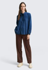 Dunkelbluse, langärmlig mit einer Fliegen-Detail am Kragen und Knopfverschluss, kombiniert mit braunen, geraden Hosen und beigen Schuhen.