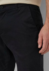 Męska prawa ręka spoczywa obok czarnych dopasowanych spodni z przednią kieszenią i zapięciem na guzik, nosząc beżową koszulę włożoną w spodnie.