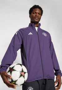 Lila Sportjacke mit schwarzen Akzenten, Reißverschluss und gesticktem Logo; das Modell hält einen weißen Fußball mit schwarzen und roten Details.