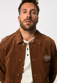 Veste en daim marron avec un col, fermeture à boutons et un patch logo brodé sur la poitrine. Portée sur un polo blanc.