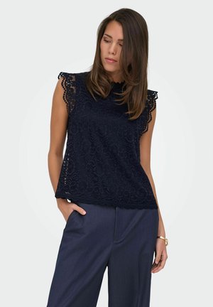 Femeie care poartă un top bleumarin fără mâneci din dantelă și pantaloni închiși la culoare, privind în jos, cu o mână în buzunar și cu o brățară aurie la încheietură.