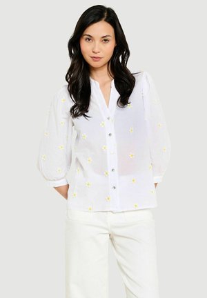 Vrouw met lang donker haar die een witte blouse met knopen draagt met kleine gele bloemembroderie en witte broek tegen een effen achtergrond.
