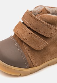 Stivaletto in suede marrone con punta in gomma grigia, due cinturini in Velcro, superficie texturizzata e una suola flessibile color nocciola.