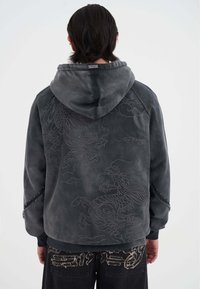 Chaqueta con capucha gris hecha de tela suave, con un intrincado bordado de dragón en la parte posterior y tachuelas decorativas en las mangas.