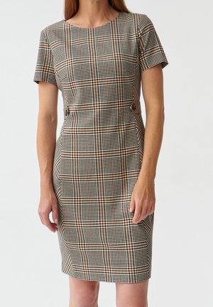 Vrouw draagt een knielange, nauwsluitende jurk met korte mouwen, in een bruin, zwart en wit geruit patroon met knoopdetails aan de taille.