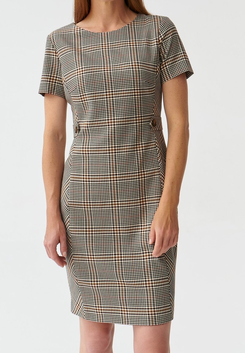 Femme portant une robe ajustée mi-longue à manches courtes, avec un motif à carreaux marron, noir et blanc, et des détails à boutons à la taille.